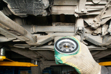 Auto mechanic holds an oil filter against the background of the bottom of a car raised on a lift....