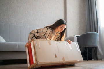 In a cozy living room, a woman sits on the floor unpacking a large cardboard box while sitting near...