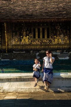 Happy mother and daughter in traditional Lao attire sitting together in front of a golden stenciled temple wall in Luang Prabang, Laos.