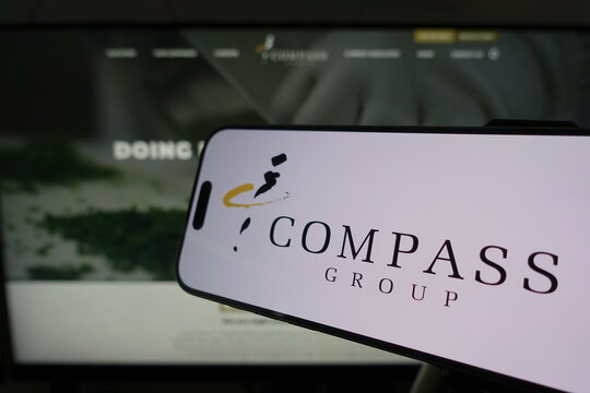 Kiel, SH, Germany March 24 2025: Smartphone displaying Compass Group logo in front of company website on a monitor illustrates modern business technology integration, showcasing digital presence