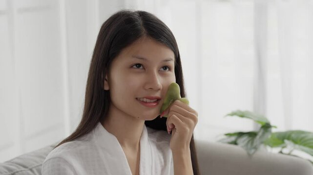 Asian woman in bathrobeses gua sha quarz lifting tool on face. Woman self massage with jade gouache scraper