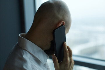 Rear view of bald businessman talking on mobile phone while looking through the office window