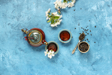 Teapot with cups, spoon of dry green tea and flowers on blue grunge background