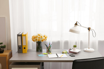 Stylish workplace with lamp, bouquet of narcissus flowers and notebook in office