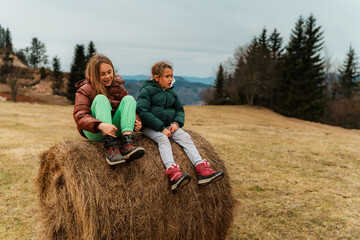 Two sisters enjoying a peaceful moment while sitting on a hay bale in a picturesque mountain meadow, surrounded by lush trees and a beautifully cloudy sky above © DusanJelicic