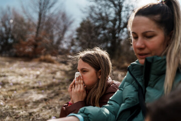 Young girl suffering from allergies or a cold, blowing her nose with a tissue while hiking outdoors with her mother, showcasing the challenges of outdoor activities with children's health