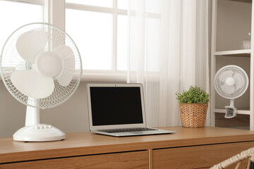 Comfortable workspace with modern laptop and electric fan on wooden desk at home office, closeup