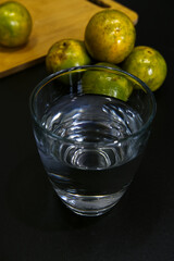 Glass of water with blurred Tangerine orange at the back with black background. Close-up water glass and fresh fruit in dark theme. Drinking, fresh fruit, room temperature water, and healthy concept.