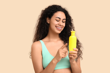 Beautiful young happy African-American woman pointing at bottle of sunscreen cream on beige background
