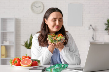 Young female nutritionist with vegetable salad video chatting at table in office