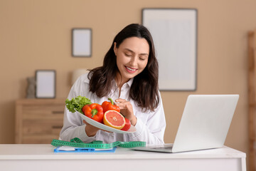 Young female nutritionist with healthy food video chatting at table in office