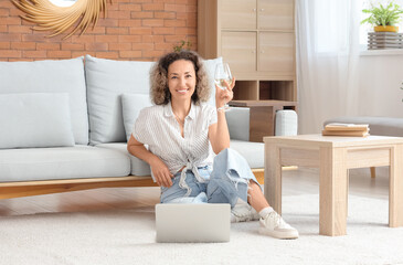 Beautiful middle-aged woman with glass of wine and laptop sitting on floor at home