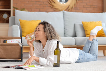 Beautiful middle-aged woman with glass and bottle of wine reading magazine at home