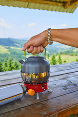 A hand lifts the lid of a metallic kettle sitting on a portable stove. The backdrop features stunning mountains under a clear blue sky, suggesting a peaceful outdoor experience.