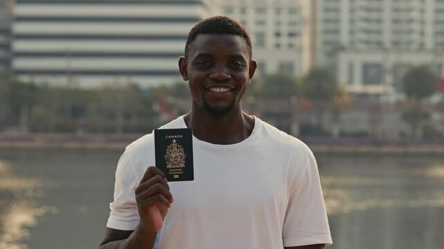 Portrait of man showing passport of Canada with cheerful smile looking at camera. African American guy celebrates acceptance into global work program on city embankment