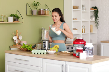 Sporty young happy Asian woman pouring milk into blender for protein shake at home