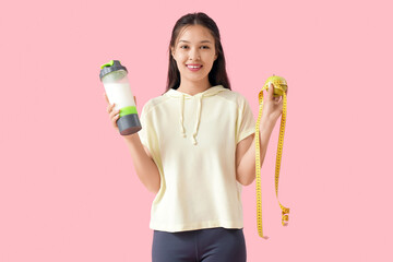 Sporty young happy woman with protein shake, measuring tape and apple on pink background