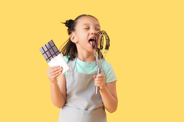 Cute little Asian girl with chocolate licking whisk on yellow background