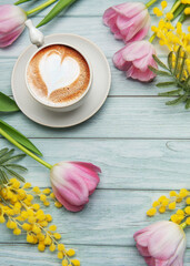 Cappuccino with heart shaped foam near pink tulips and mimosa flowers on light blue wooden table