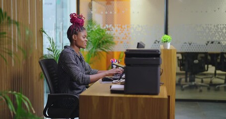 African young adult gen z woman lady sitting chair desk table use computer look screen do work task typing email check data at job office expert staff busy female girl attend phone calls talk day time