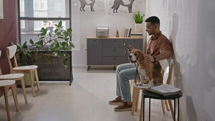 Young man sitting in veterinary clinic waiting room with beagle on leash, using smartphone while waiting for medical appointment