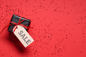 Gift box with ribbon, label and confetti on red background. Black Friday sale. Top view