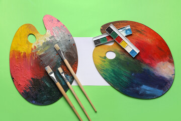 Artist brushes, palettes, sheet of paper and paints on green background