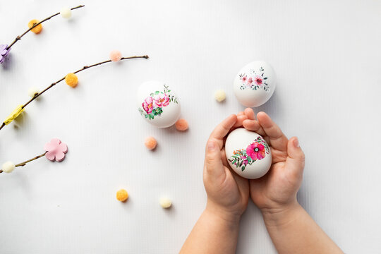 Hands holding three white eggs with floral designs, surrounded by colorful pom-poms on a white surface. Perfect for Easter or spring.