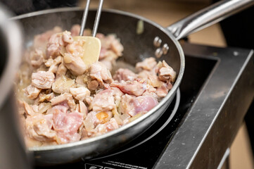 A close-up view of diced chicken being sautéed in a silver frying pan. Steam rises as the meat cooks, alongside chopped onions, in a modern kitchen setting.