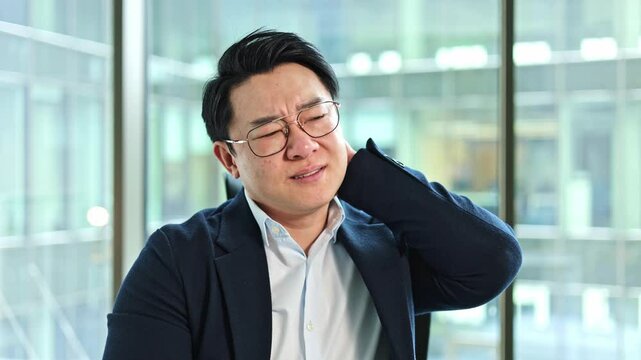 Stressed Korean businessman has neck pain in a modern office. Exhausted Asian man massages his neck. Male entrepreneur in glasses feels uncomfortable at work.