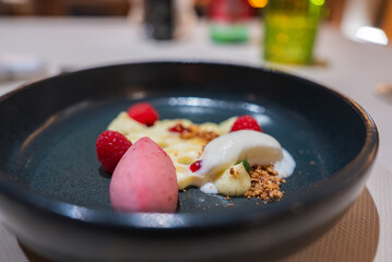 A dessert featuring pink sorbet, raspberries, custard, white mousse, and nuts in a dark bowl. Blurred dining table setup with colorful glassware visible.
