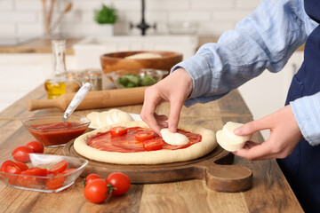 Woman putting mozzarella cheese on dough for pizza near table in kitchen