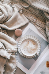 Cup of frothy coffee on a white scalloped saucer, styled with neutral textiles, a macaron, and a fashion magazine. Cozy and elegant aesthetic in soft beige tones