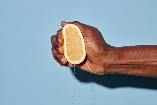 Close-up of hand squeezing fresh lemon half with juice dripping. Background is plain light blue creating strong contrast with the hand and lemon