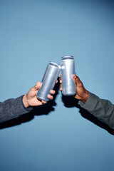 Close-up of two people's hands holding and clinking aluminum cans, set against a blue background,...