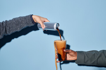 Hand pouring coffee from silver can into clear glass cup held by another hand against simple blue...