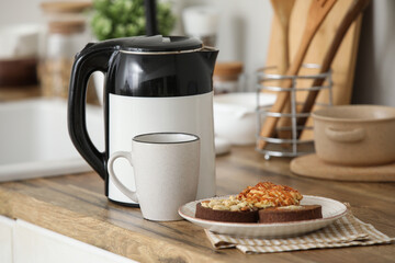 Modern electric kettle with cup of hot tea and sweet cakes on counter in kitchen