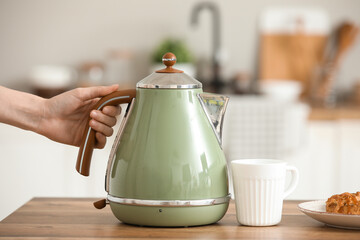 Female hand with modern green electric kettle, cup of hot tea and sweet cake on wooden table in...