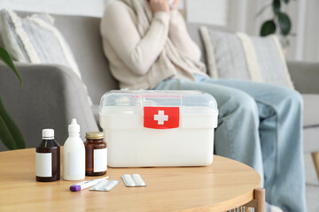 Sick woman sitting on sofa at home, focus on table with pills, bottles of medicine and first aid kit