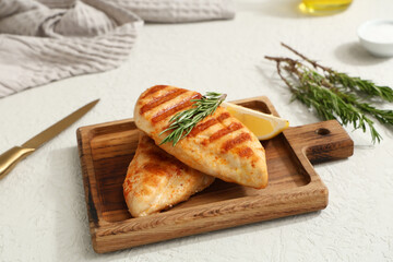 Wooden board with tasty grilled chicken breast, rosemary and lemon slice on white background
