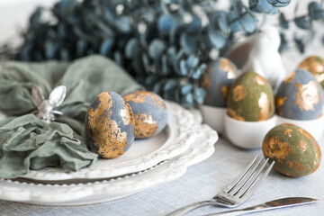 The banner. Table setting. A plate with a rabbit and a napkin, Easter eggs with gilding and a branch of eucalyptus on a linen tablecloth. The concept of a bright Easter holiday.