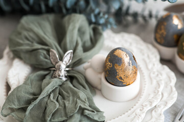 The banner. Table setting. A plate with a rabbit and a napkin, Easter eggs with gilding and a branch of eucalyptus on a linen tablecloth. The concept of a bright Easter holiday.
