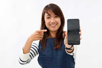 Fototapeta premium Young asian barista woman wearing apron pointing finger at mobile screen,showing app and smiling, standing over white background