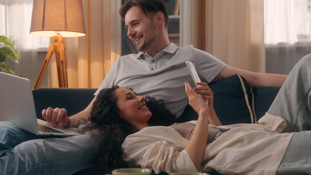 Gadget addict family internet addiction couple Caucasian man freelancer guy male using laptop computer Hispanic woman user Latina girl lady together use mobile phone smartphone at home couch relaxing