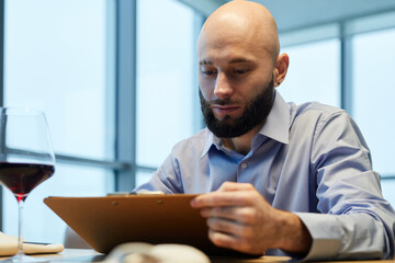 Young man sitting at table in the restaurant with glass of wine and reading menu