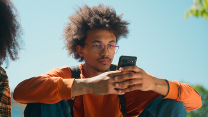 Two African American students friends woman girl female man male guy together gadget addict using mobile phones scrolling smartphones social media sit in city outdoor of college high school university