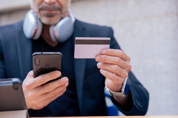 Unrecognizable caucasian man with headphones smiling while using smartphone and holding a credit card outdoors. Concept of online shopping, digital payment, modern lifestyle, and financial technology.