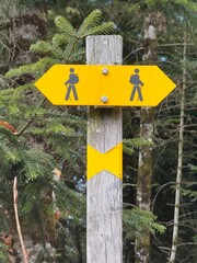A yellow hiking trail signpost in a forest, mounted on a wooden pole, points in two opposite directions. The sign features two black hiker icons with backpacks and trekking poles. Green trees surround