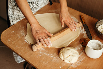Female hands rolling out dough on a wooden table with a rolling pin. Flour, kitchen ingredients and utensils around. Process of making homemade pastries