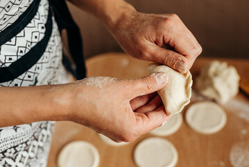 Female hands are molding dumplings on a kitchen table sprinkled with flour. Round dough blanks are cut out nearby. Home cooking, preparation of traditional dishes